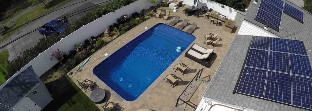 Residential in-ground swimming pool with adjacent house or structure featuring rooftop solar panels used for pool heating or home energy, surrounded by clean decking and landscaped backyard.