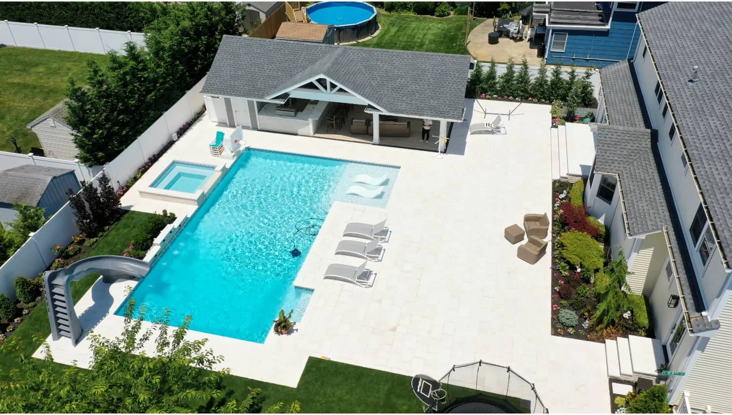 Aerial view of backyard with custom inground pool, hot tub, slide, lounge chairs, patio, trampoline, and above-ground pool