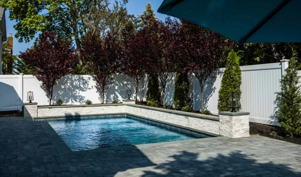 Rectangular in-ground swimming pool surrounded by stone pavers, with trees, shrubs, and a white privacy fence in the background. Expert inground pool installation by a trusted pool company. Part of a blue umbrella is visible in the top right corner.