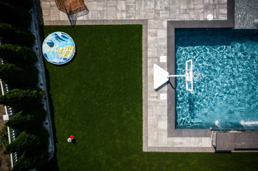 Aerial view of a backyard featuring a rectangular custom pool by a trusted pool company, with green grass, a round float, a basketball hoop near the inground pool, and a small red and white ball on the lawn.
