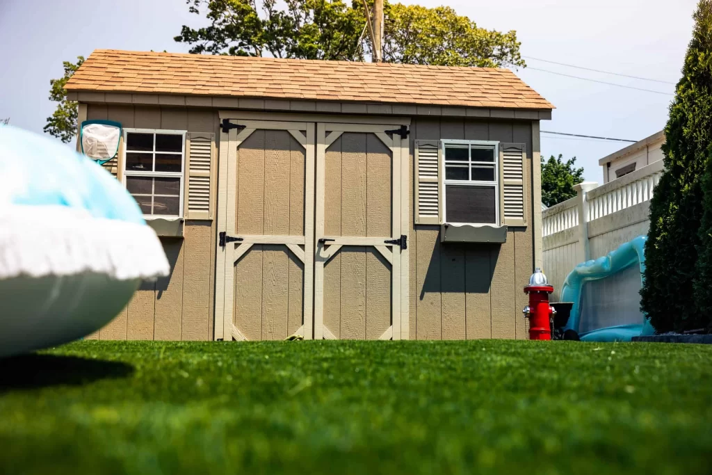A tan garden shed with white shutters stands on green artificial grass, flanked by a red fire hydrant and a blue inflatable pool, suggesting recent pool installation. Trees and a white fence are in the background, with a blurred object in the foreground.