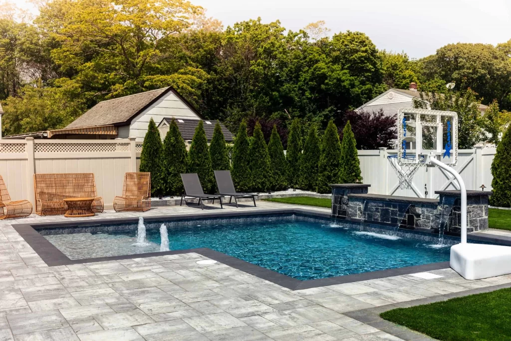 A backyard inground pool installation features water fountains, a basketball hoop, lounge chairs, and wicker chairs. Trees, a white fence, and a small house in the background create a private and inviting outdoor space.