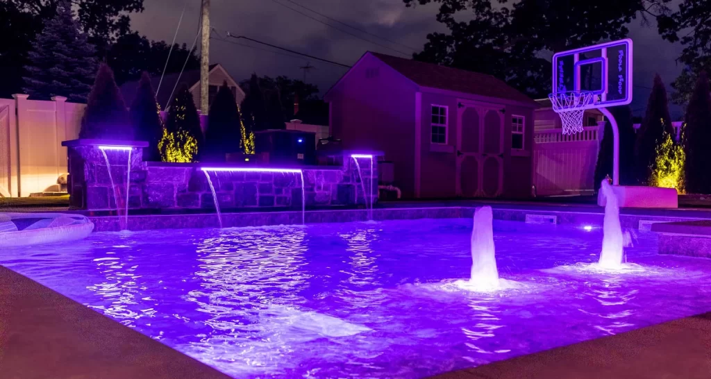 Backyard swimming pool with purple LED lighting at night, water features, basketball hoop, stone wall, illuminated landscaping, and a shed in the background in Long Island