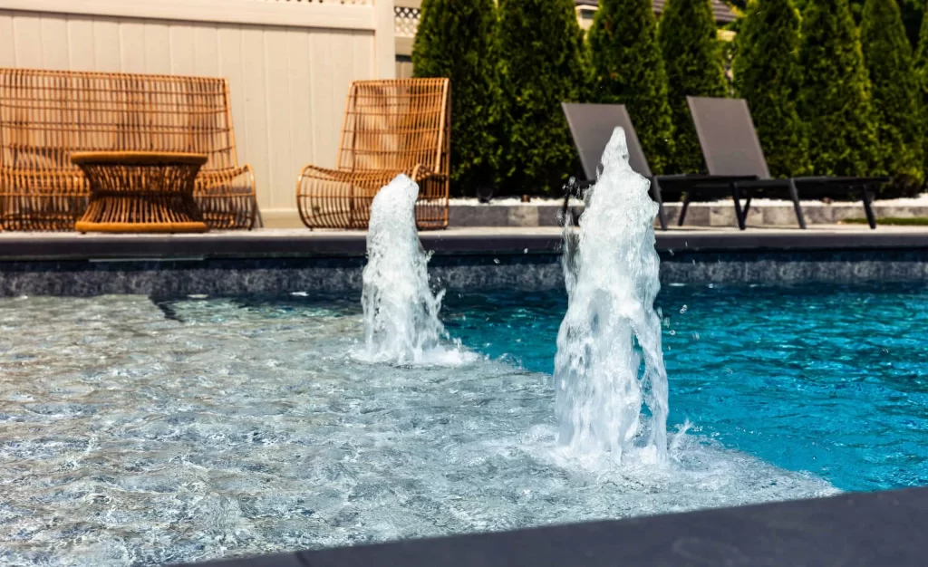Water jets spray upward in a backyard pool, showcasing expert inground pool installation. Wicker furniture and lounge chairs are arranged on the deck, surrounded by green trees and a privacy fence in the background.