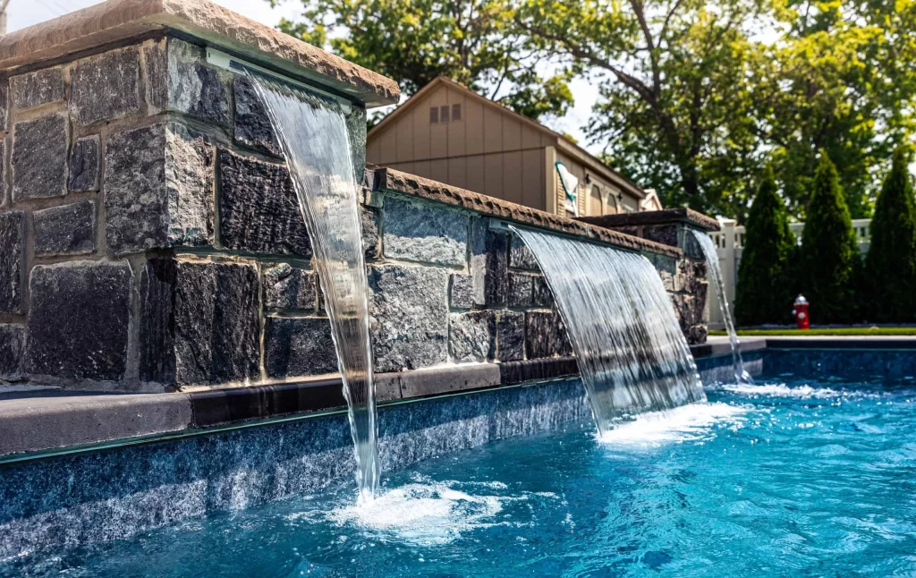 Water cascades from two stone spouts into a clear blue swimming pool, showcasing expert pool installation, with trees, a house, and a fence visible in the background on a sunny day.