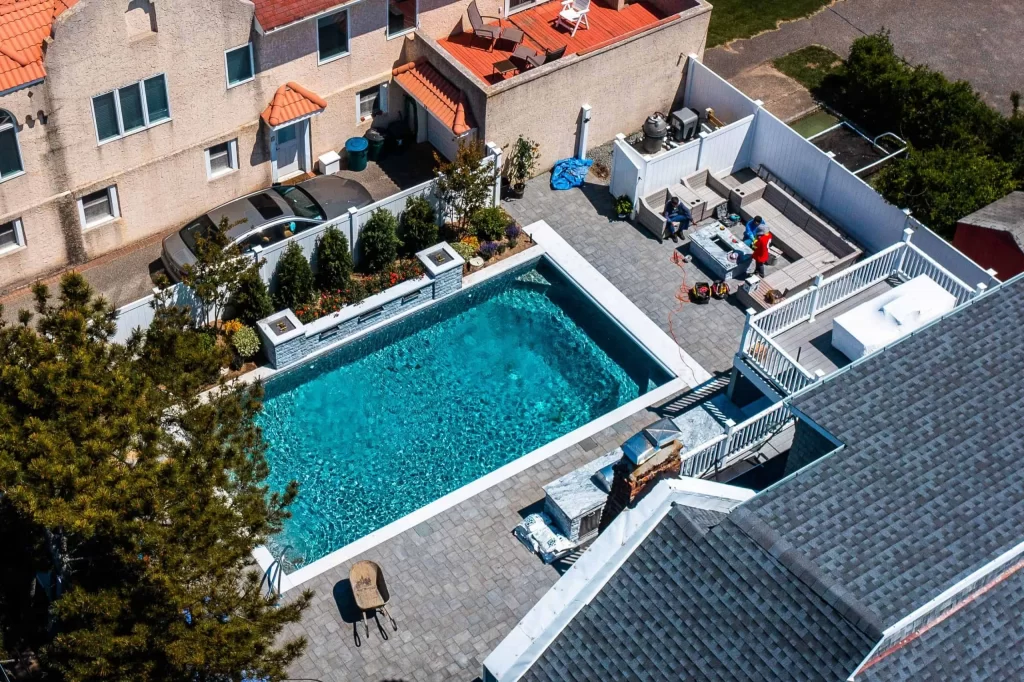 Aerial view of a backyard with a rectangular swimming pool by a top Pool Company, patio furniture, people gathered near a grill, parked cars, and surrounding houses with red tile and gray shingle roofs.