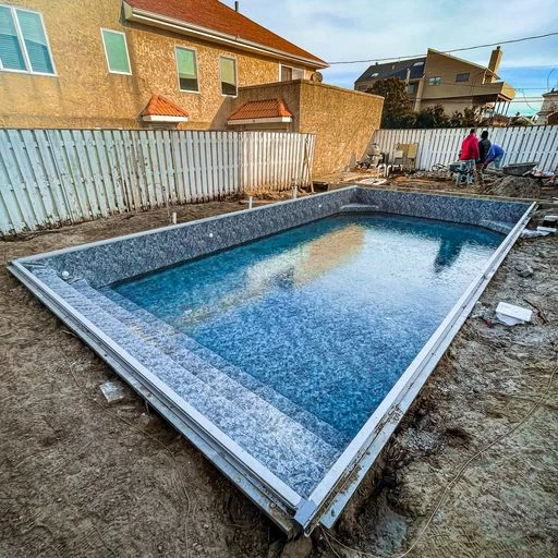 A rectangular inground pool installation is underway in a backyard, partially filled with water. Construction materials and workers surround the site, with houses and a white fence in the background—highlighting expert pool services.