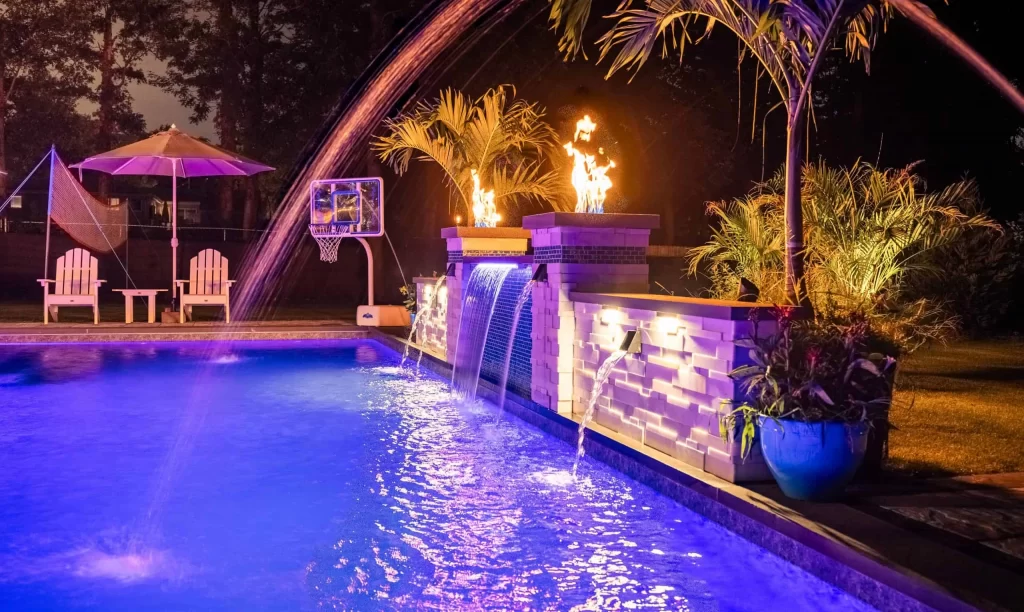 Backyard swimming pool at night with illuminated waterfall feature, basketball post, and poolside chairs showcasing a blend of recreation and relaxation in a custom pool installation