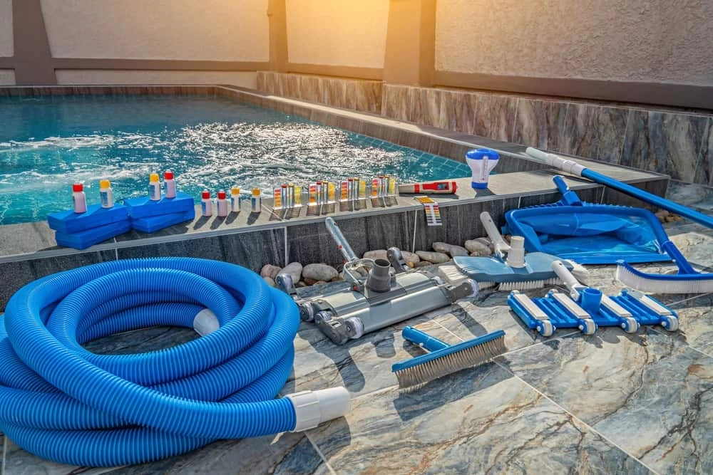 Various pool cleaning equipment, test kits, hoses, brushes, and nets are neatly arranged by the edge of a swimming pool installed by a trusted pool company, with sparkling water visible in the background.
