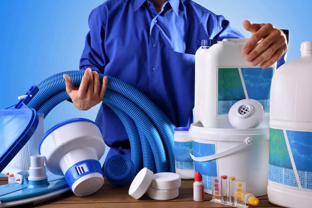 A person in a blue shirt holds pool maintenance supplies, including hoses, containers, chlorine tablets, and test kits&mdash;perfect for anyone considering inground pool installation or seeking advice from a trusted pool company.
