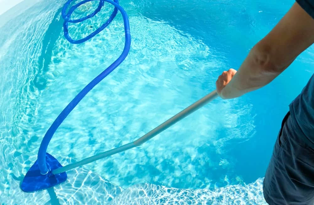 A person cleaning a swimming pool with a long-handled vacuum and blue hose trailing in the clear water, showcasing the sparkle of custom pools after expert inground pool installation by a professional pool company.