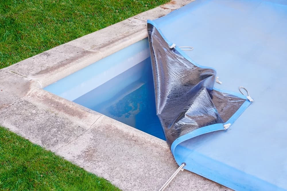 A rectangular swimming pool is partially covered with a blue protective cover, pulled back at one corner to reveal clear water. The pool, surrounded by stone paving and green grass, showcases quality inground pool installation and custom pools expertise.
