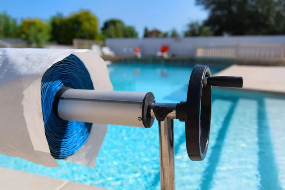 A close-up of a pool cover roller with a blue cover rolled up, positioned at the edge of a clear blue swimming pool on a sunny day—perfect inspiration for custom pools or inground pool installation. Trees and outdoor furniture complete the scene.