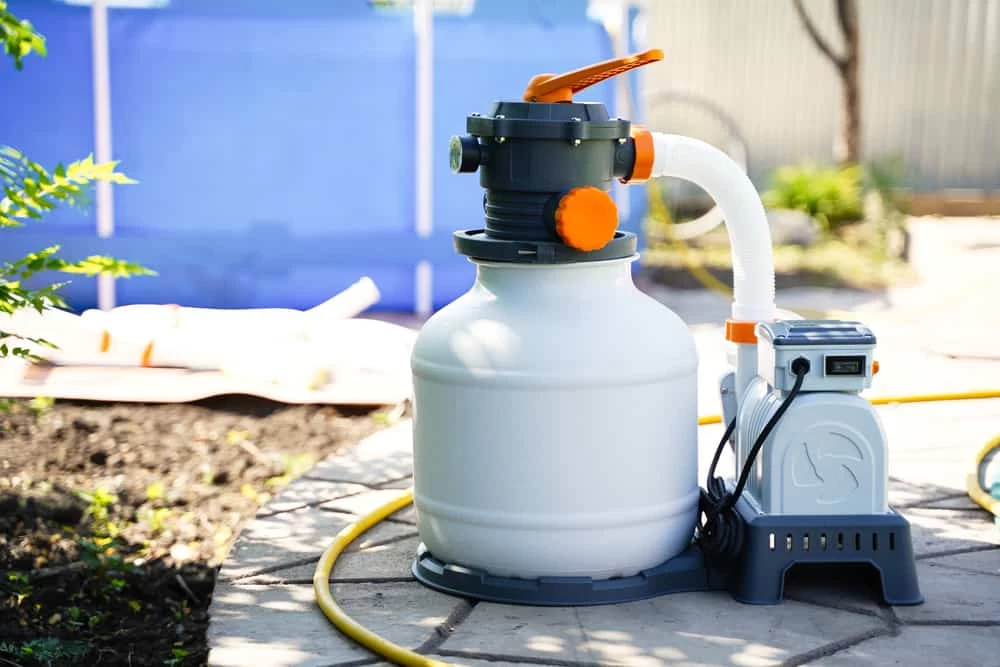 A white pool filter pump with hoses and an orange handle is set up on a stone patio near a blue above-ground pool in a backyard—ideal for those considering professional pool services to keep their setup running smoothly.
