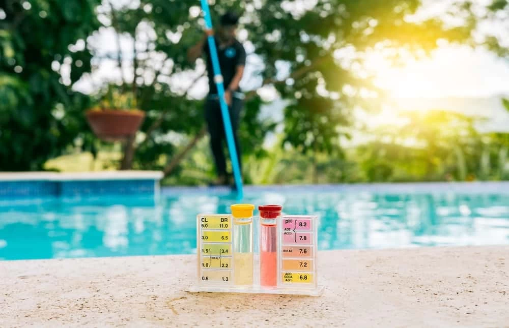 Pool water test kit with colored vials and charts sits on a poolside surface. In the blurred background, a person cleans the pool with a pole—part of routine pool services—surrounded by greenery and sunlight.