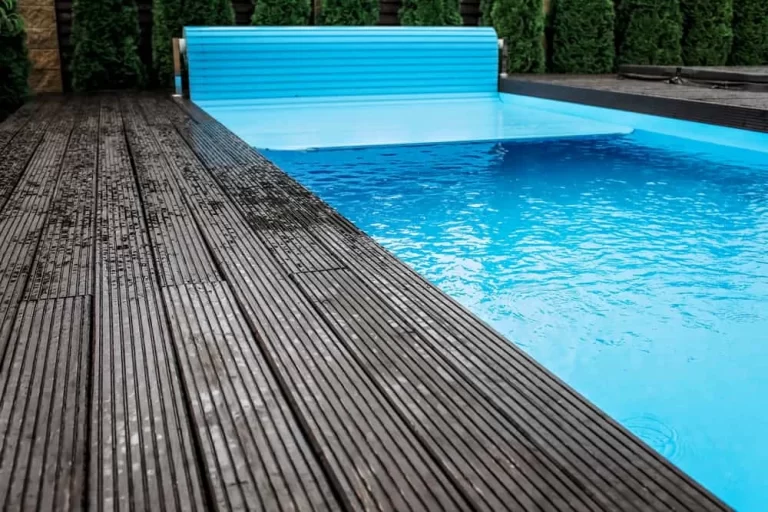 A swimming pool with a bright blue automatic pool cover partially unrolled, surrounded by wet, dark wooden decking and green shrubs—an inviting scene showcasing professional inground pool installation.