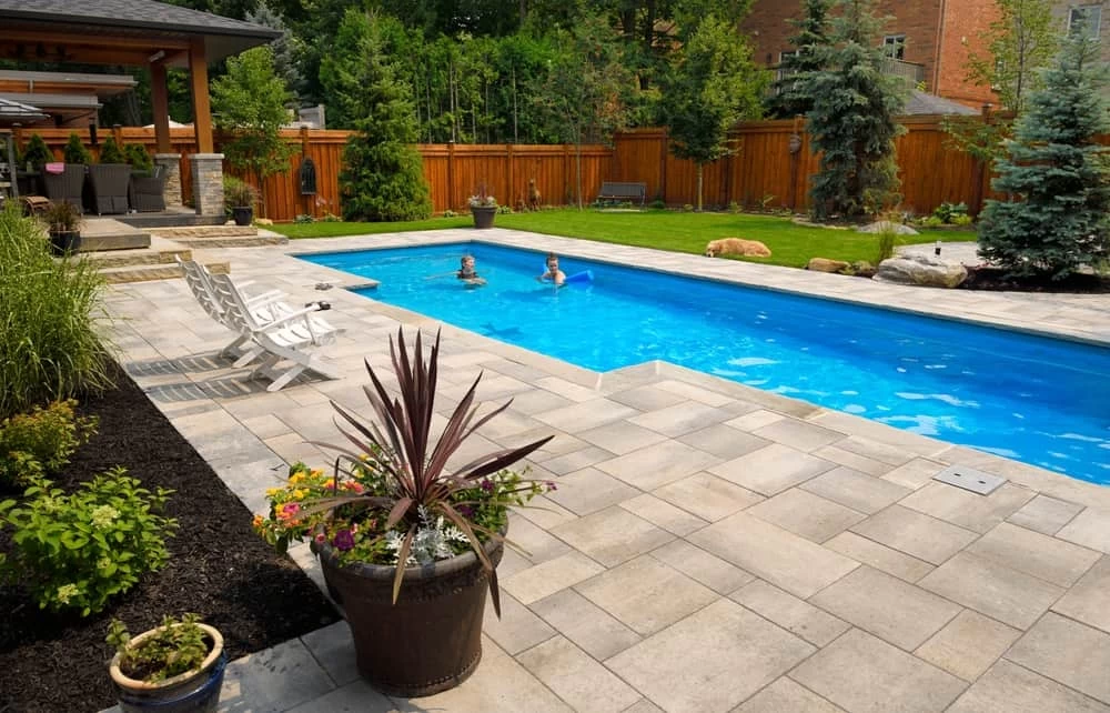 A backyard with a rectangular inground pool installation, two people swimming, lounge chairs on a stone patio, potted plants, green lawn, and a wooden fence surrounded by trees and shrubs.