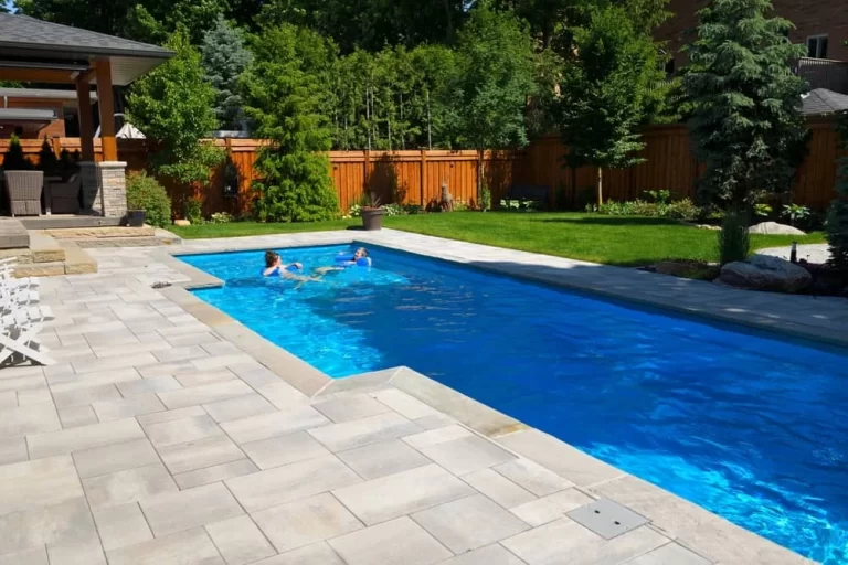 Two people swim in a custom pool surrounded by stone tiles, lush grass, trees, and a wooden fence. On the left, a covered patio with outdoor furniture overlooks this inviting inground pool installation.