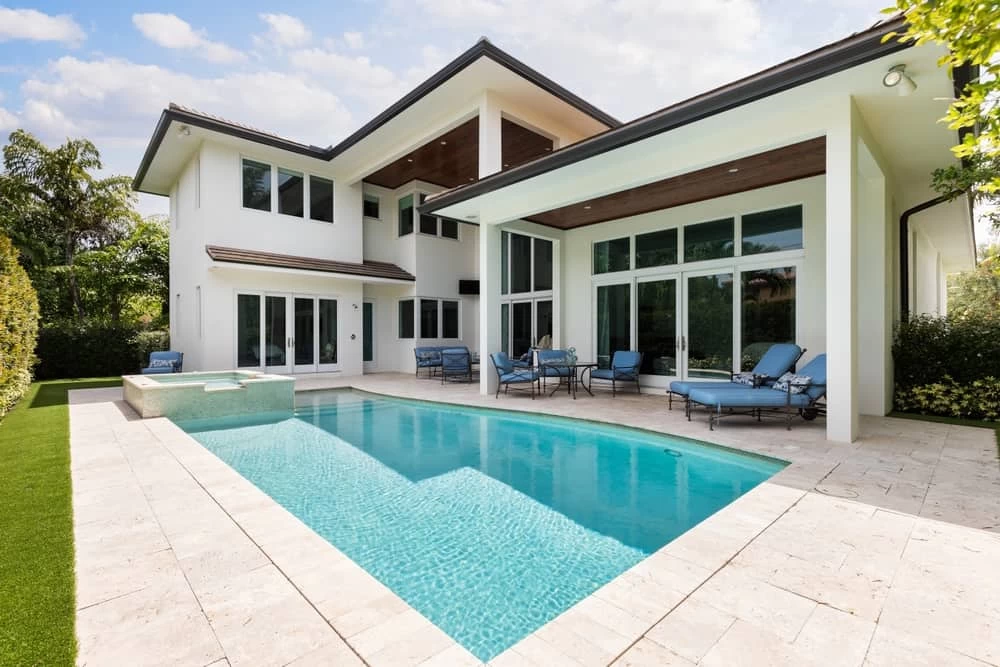 Modern two-story house with large windows, outdoor patio furniture, and a swimming pool with a built-in spa—expertly crafted through custom pools and quality pool installation—surrounded by greenery and a clear sky above.