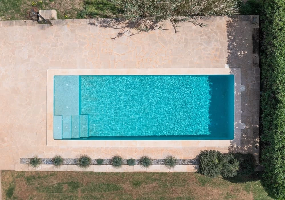 A rectangular swimming pool with clear blue water is surrounded by a stone patio and lush greenery, viewed from above. Steps lead into the pool from one end—perfect inspiration for custom pools designed by a trusted pool company.