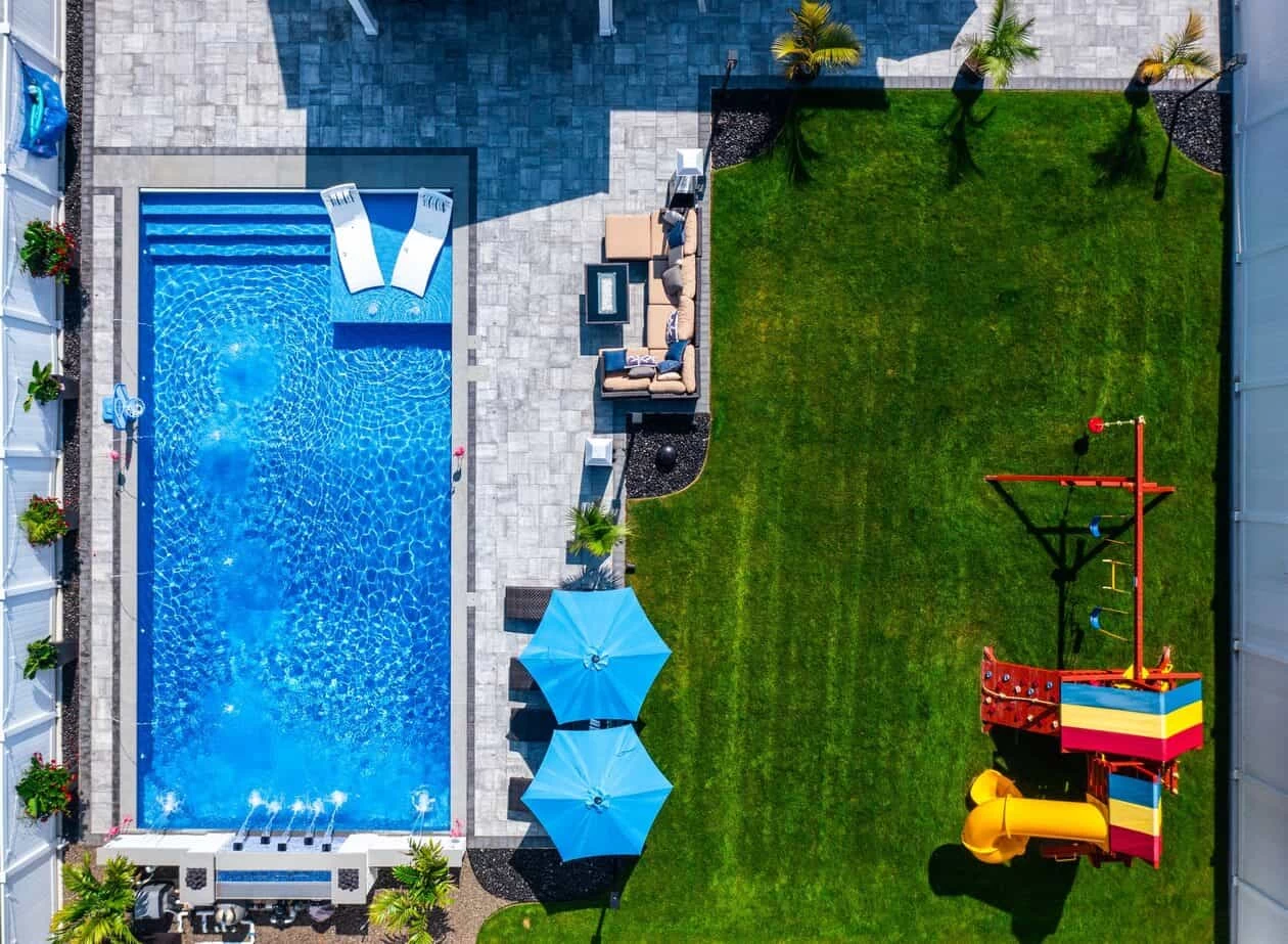 Aerial view of a backyard with a custom pool, lounge chairs, patio seating, blue umbrellas, and a green lawn with a colorful playset featuring slides and swings.