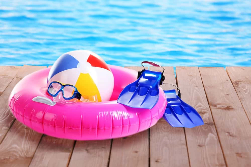 A pink inflatable swim ring, a colorful beach ball, blue swim fins, and a snorkel mask rest on a wooden deck by a bright blue swimming pool—perfect inspiration for your own custom pool from a trusted pool company.