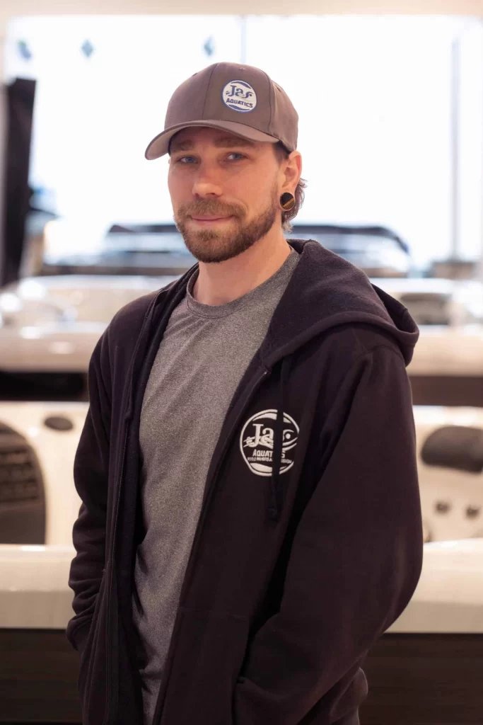 A man with a beard wearing a gray cap, gray t-shirt, and dark hoodie stands indoors, smiling slightly.