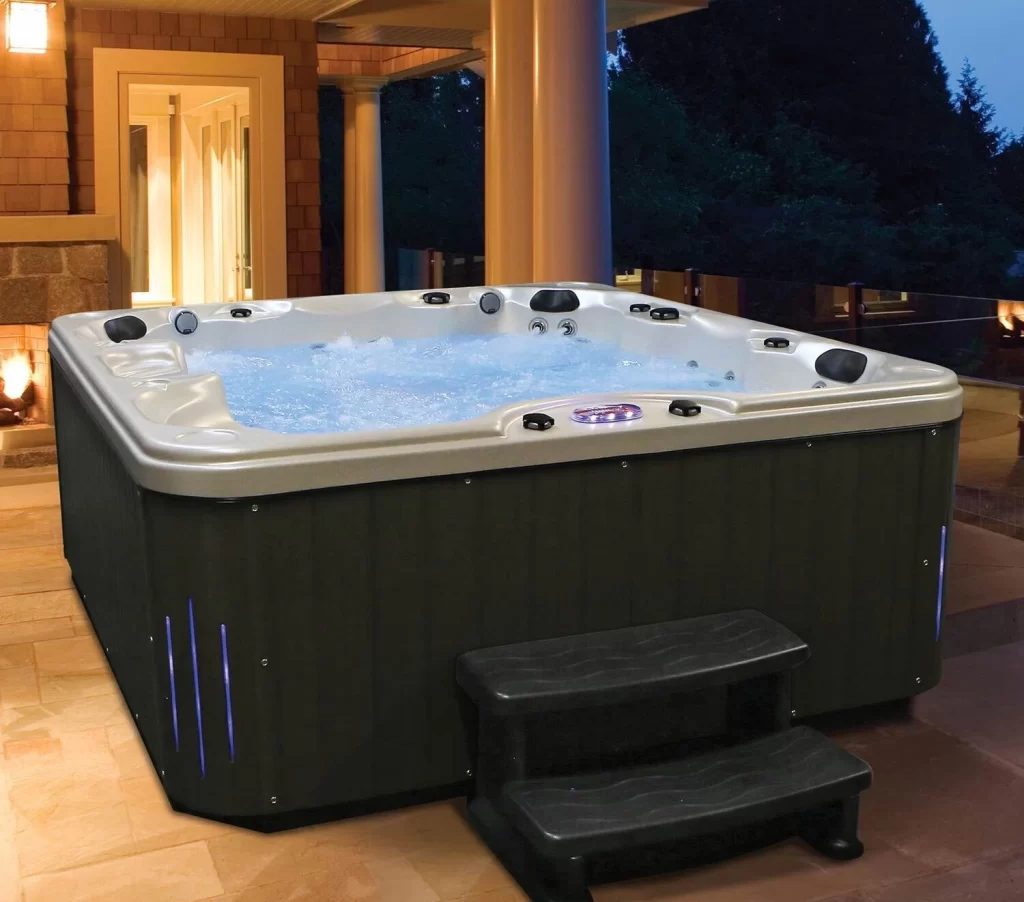 Outdoor hot tub with bubbling water on tiled patio next to house, black steps leading up, warm evening lighting, and trees in background—ideal complement to custom pools by Long Island pool company