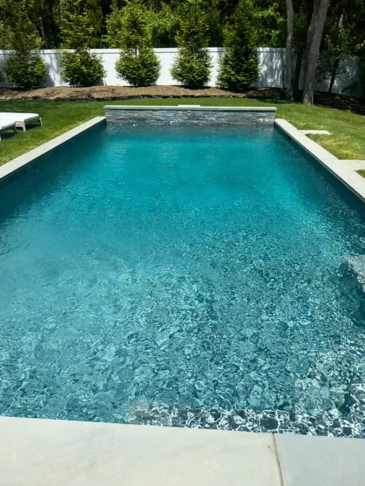 A rectangular outdoor swimming pool with clear blue water, surrounded by a stone deck and green grass. Lounge chairs are visible on the left side, and tall green trees line the background.