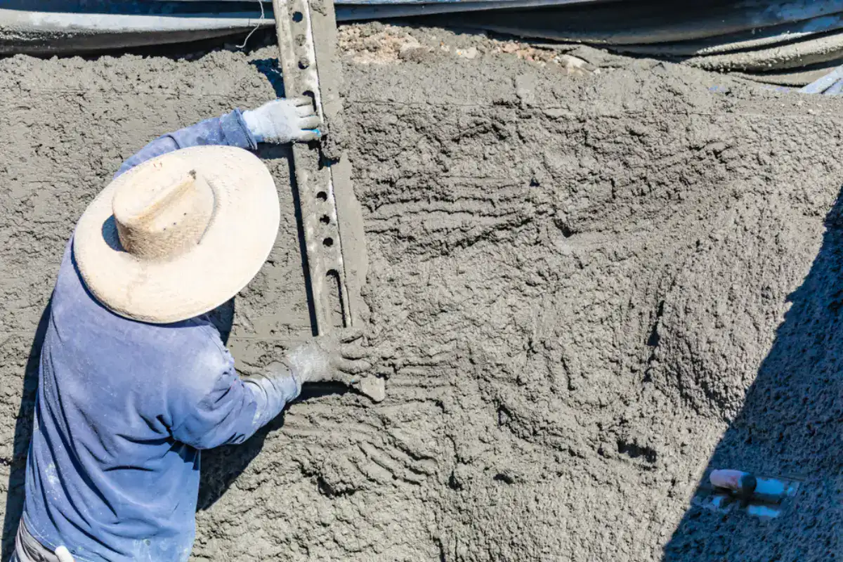 A construction worker wearing a straw hat and gloves smooths wet concrete with a metal leveling tool at a construction site.