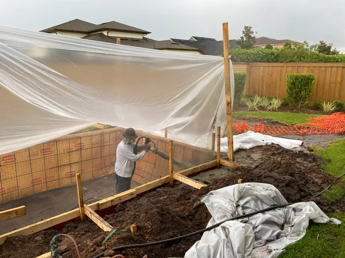 A worker uses a power tool inside a partially built structure, covered by a large plastic sheet, with muddy ground and construction materials around in a residential backyard.