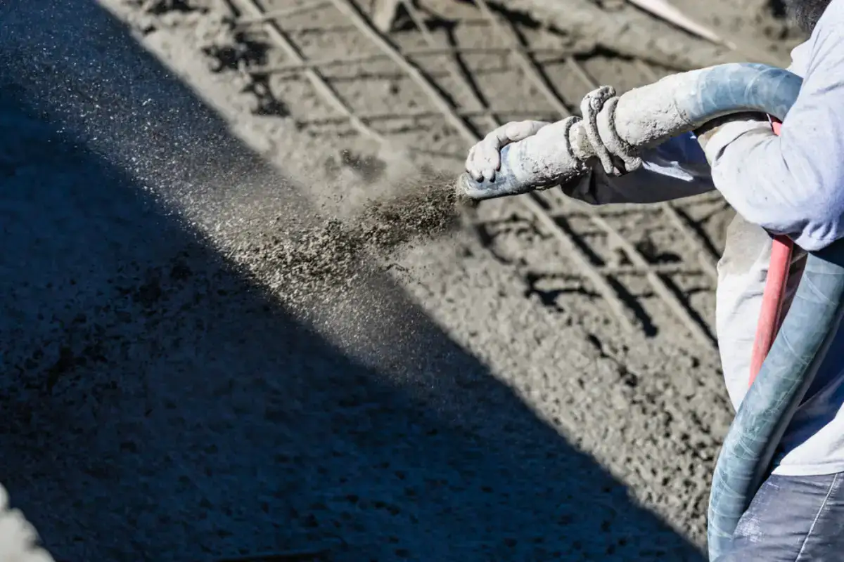 A construction worker sprays wet concrete from a hose onto a surface, with steel rebar visible underneath and shadows cast across the area.