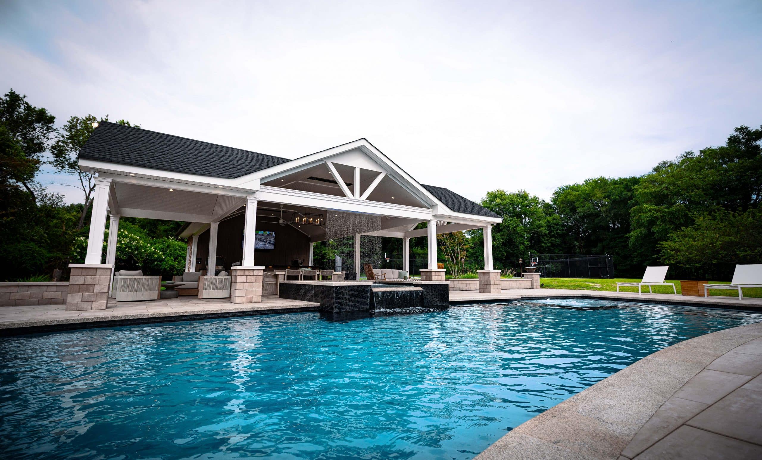 A modern outdoor pavilion with a covered seating area overlooks a large, rectangular swimming pool with clear blue water. Surrounded by trees, greenery, and lounge chairs, this setting is perfect for showcasing custom pool photos.