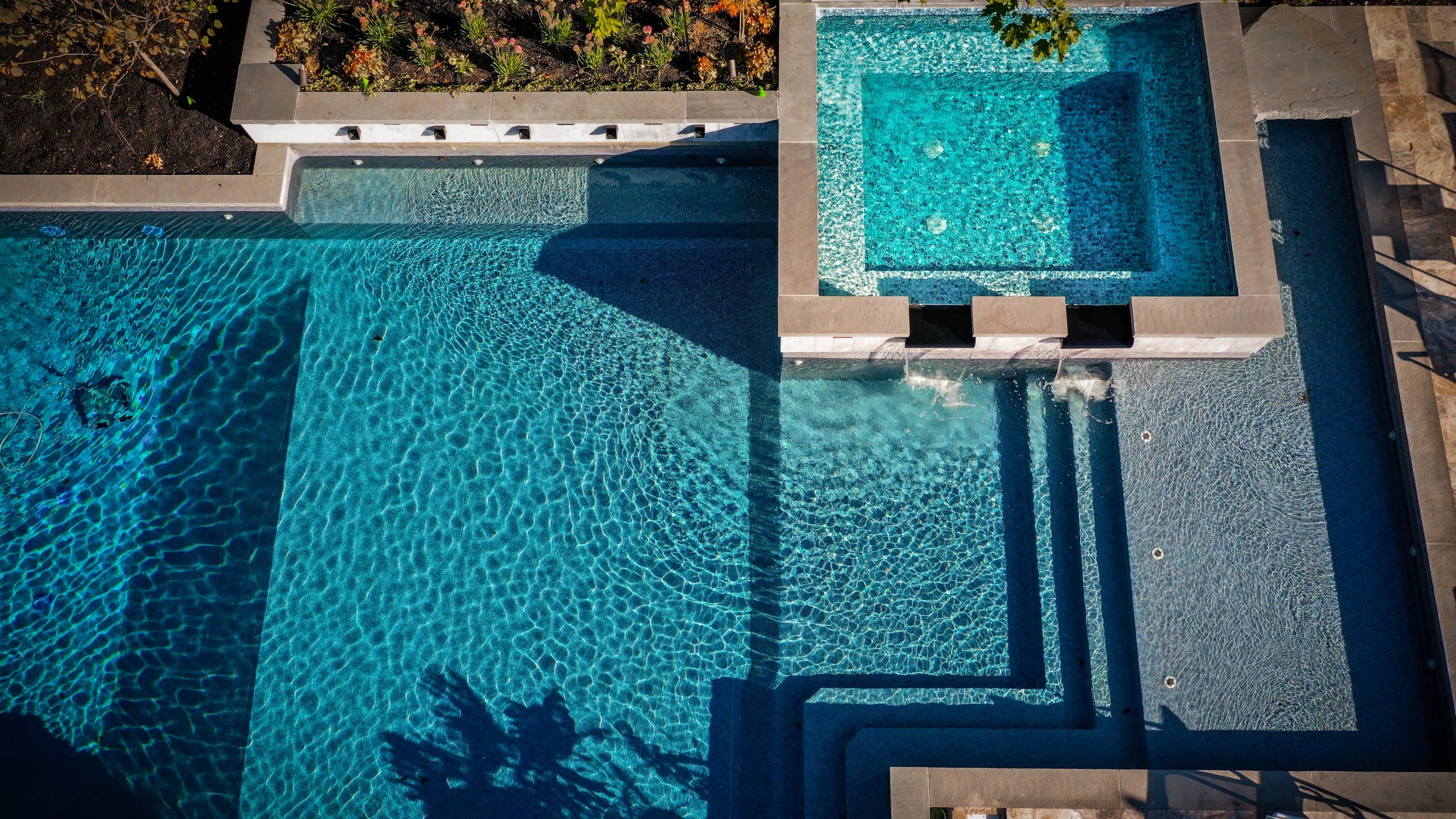 Aerial view of a modern swimming pool with a connected square hot tub, clear blue water, steps leading in, small jets, and landscaped plants around the edge&mdash;perfect inspiration for custom pool photos.