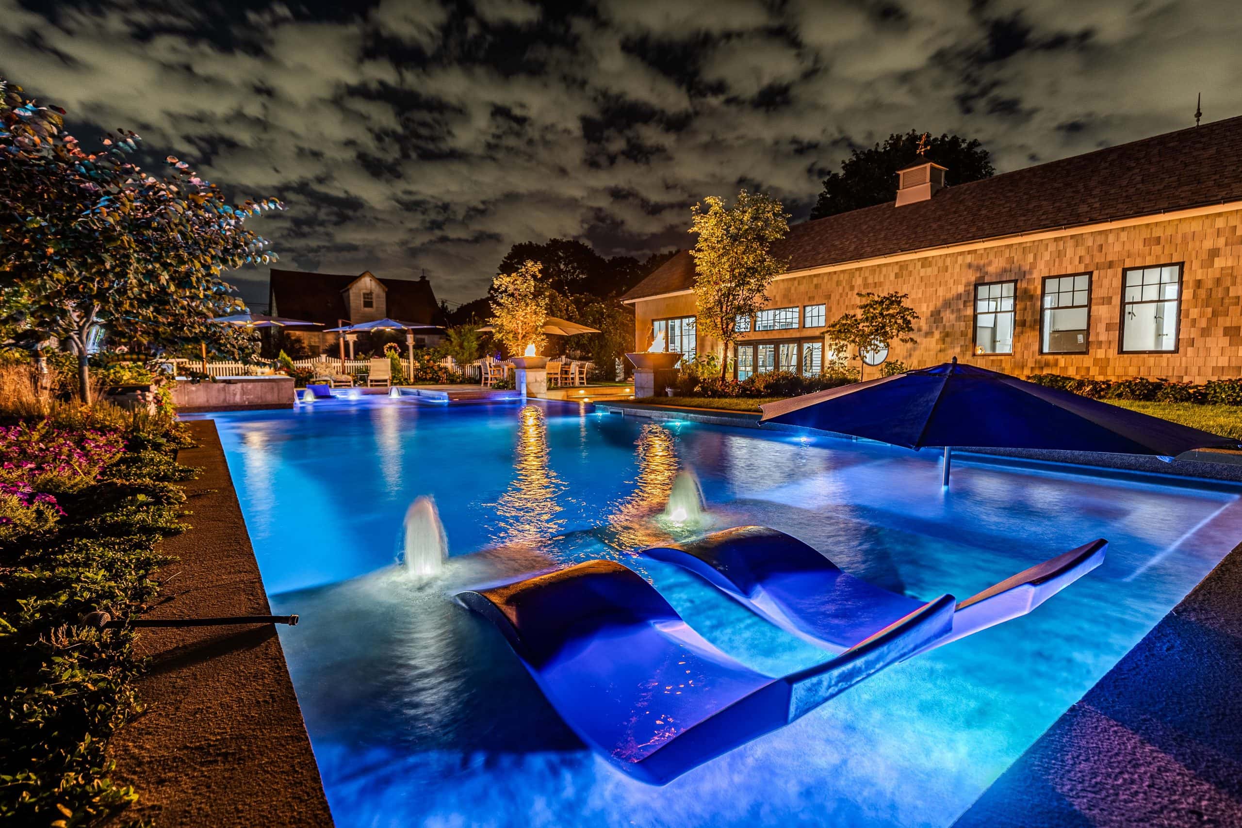 A swimming pool at night glows with blue lights, featuring two blue lounge chairs partially submerged in water. Custom pool photos capture the fountains, trees, and nearby buildings silhouetted under a cloudy sky.