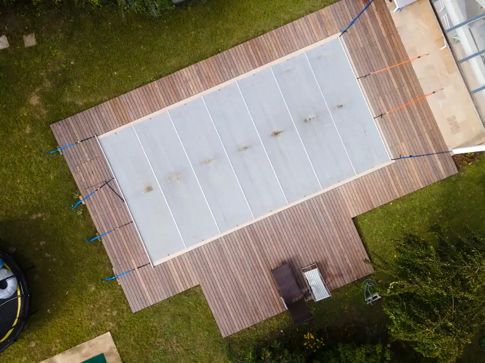 Aerial view of a rectangular swimming pool covered with a white pool cover, surrounded by a wooden deck and green grass. A bench and small table sit on the deck, highlighting the benefits of professional pool closing services.