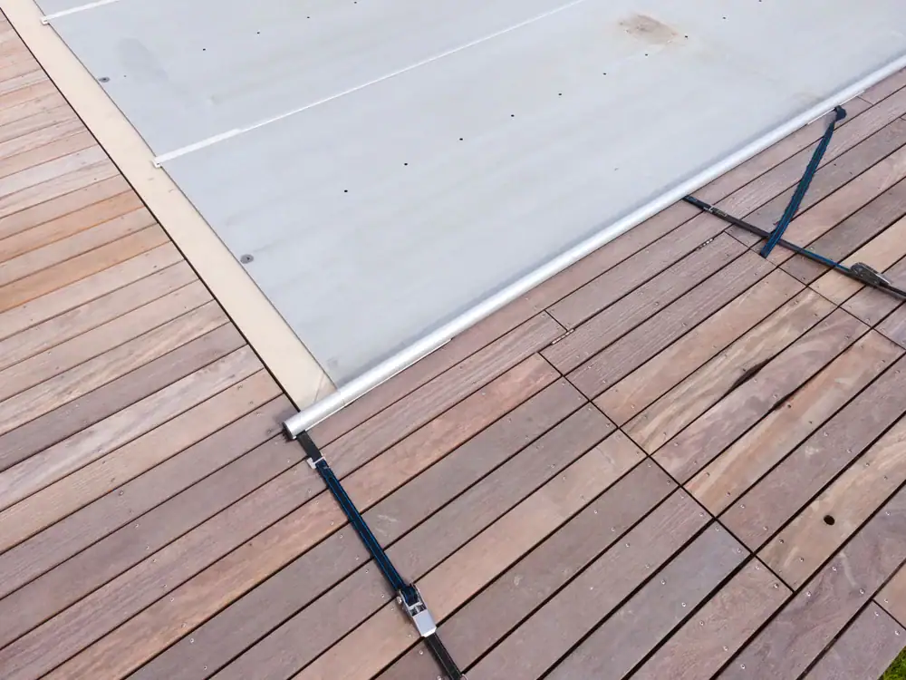 A close-up view of a wooden deck with a partially extended gray retractable cover, supported by metal rods attached to the decking—perfect for those considering pool closing services.