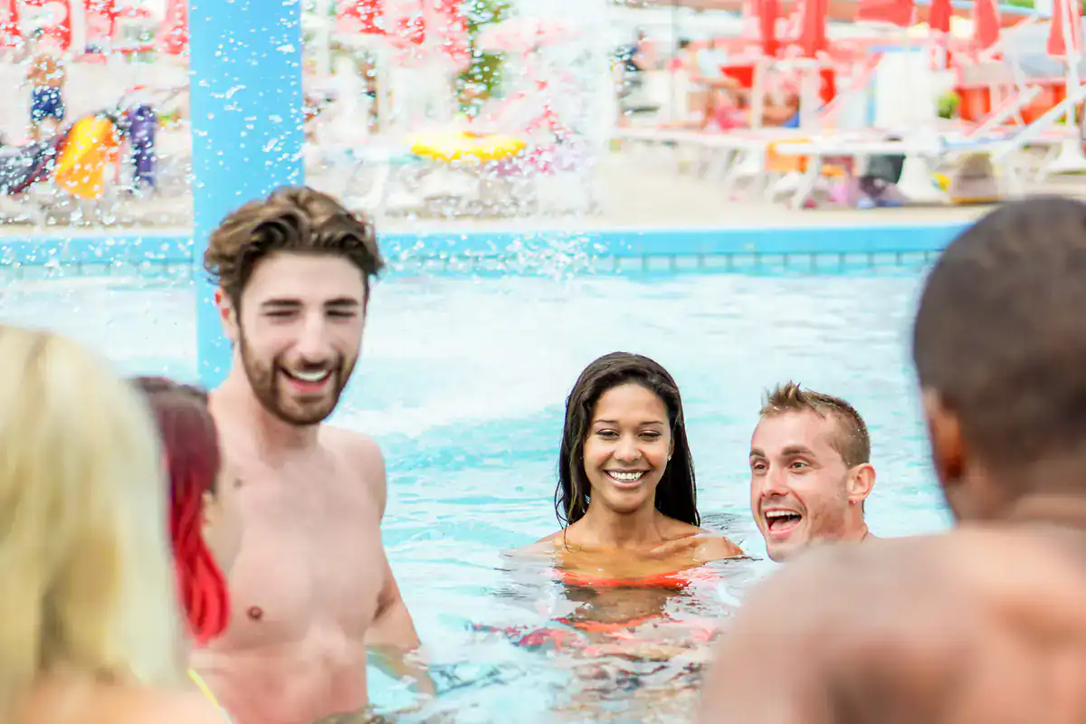 A group of young adults smiling and talking while standing together in a swimming pool, with splashing water and sun loungers in the background.