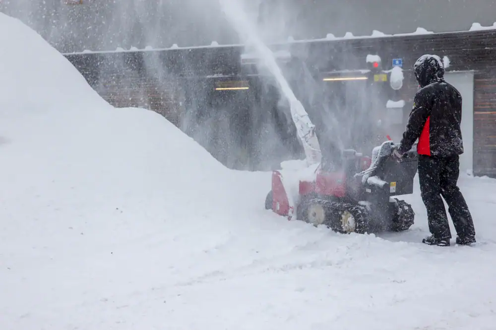 A person in winter clothing operates a red snow blower, clearing a thick layer of snow near a large snowbank in front of a building during heavy snowfall.