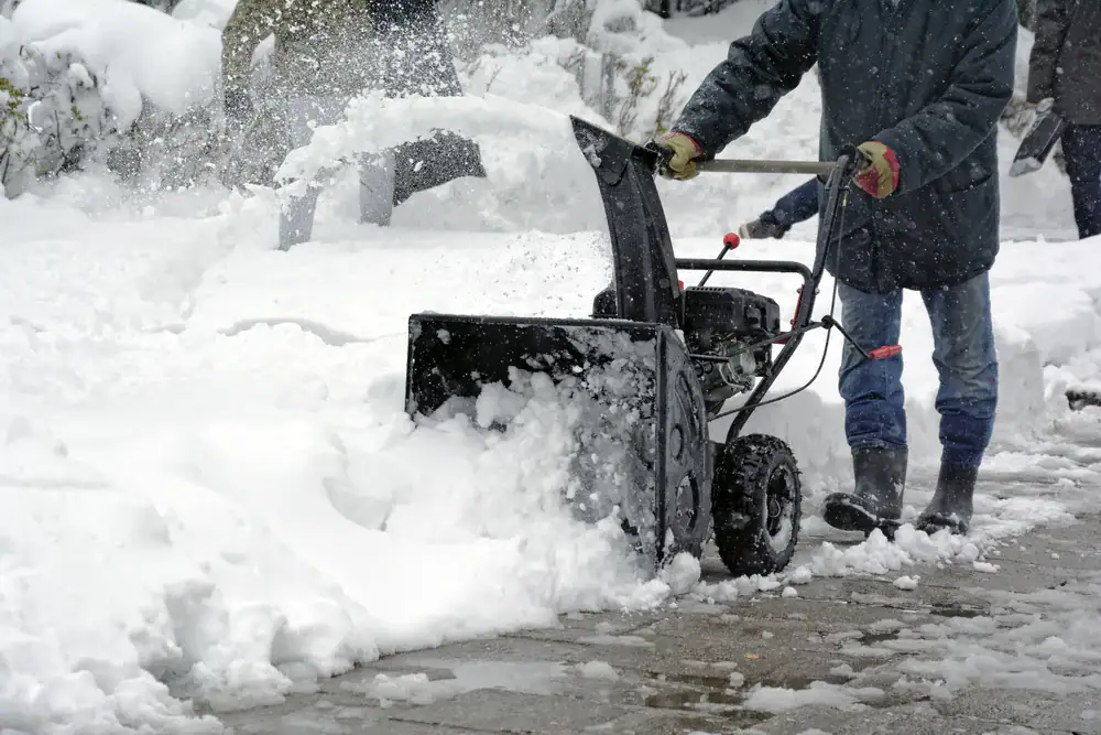 A person uses a snow blower to clear a thick layer of snow from a sidewalk. Snow is seen being discharged to the side, and the person is dressed in winter clothing and boots.