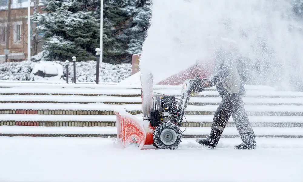 A person operates a snow blower on a snowy sidewalk, clearing a thick layer of snow as flakes swirl around them. Steps and trees covered in snow are visible in the background.