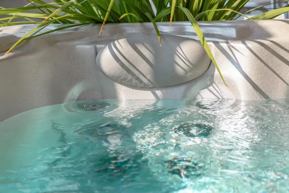 Close-up of a bubbling hot tub filled with clear water, with a built-in headrest and green plant leaves hanging over the edge, casting shadows on the tub&rsquo;s surface.