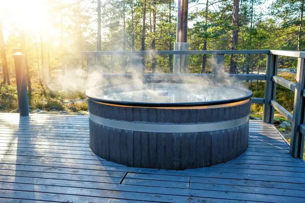 A steaming outdoor hot tub sits on a wooden deck surrounded by trees, with sunlight streaming through the forest in the background.