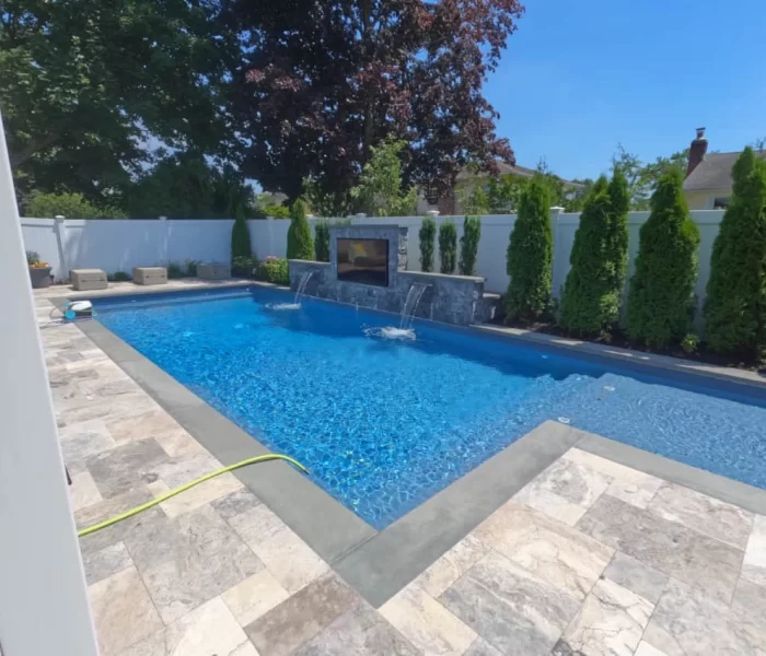 A backyard swimming pool with blue water, a waterfall feature, and a stone deck by a top Pool Company. Tall green shrubs line the white privacy fence, and a yellow garden hose is on the patio. It's a sunny day with clear blue skies.